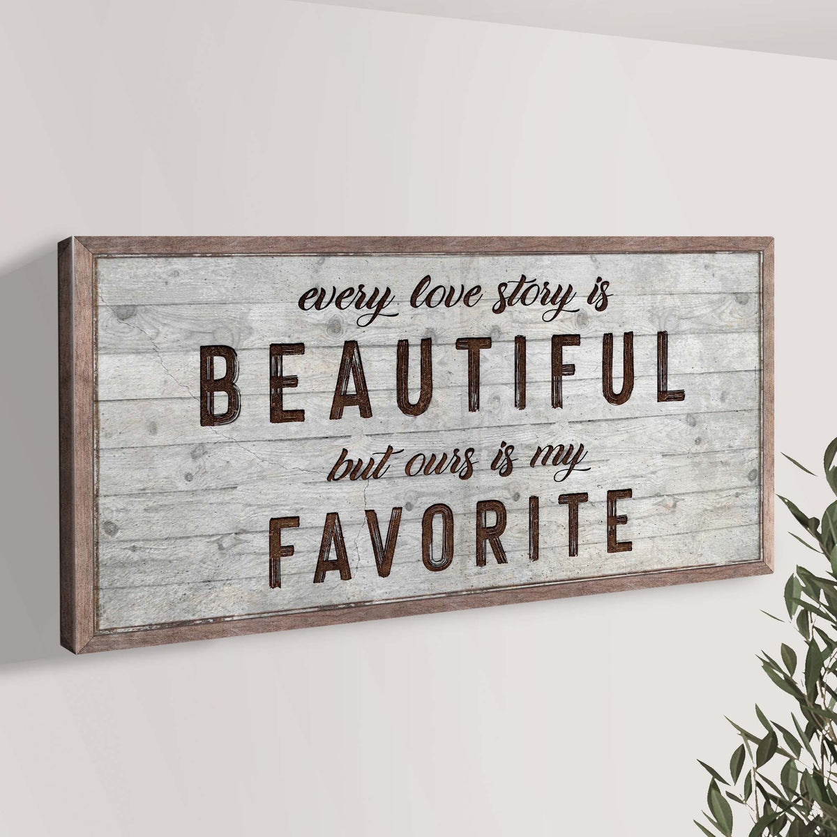 Wide love story canvas sign with dark lettering on gray planks, angled on a pale wall beside olive branches