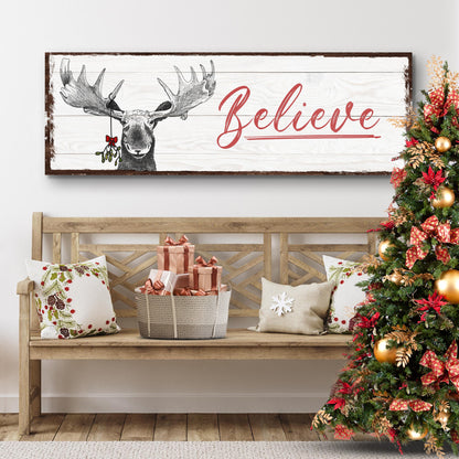 Believe canvas sign with a moose wearing holly on white wood-look planks, above a bench beside a Christmas tree