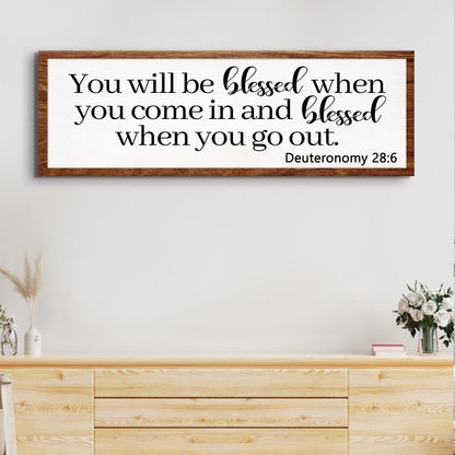 Long white canvas sign with a brown wood-look edge reading You will be blessed when you come in, Deuteronomy 28:6