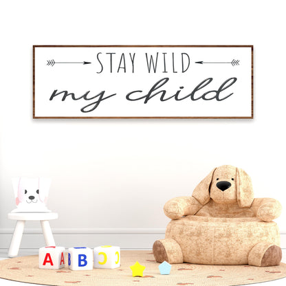 Stay Wild My Child canvas sign with two arrows on white, above a plush dog chair and toy blocks, nursery wall decor