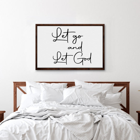 White canvas sign with fine black script reading Let go and Let God in a dark wood-look border, above a white bed