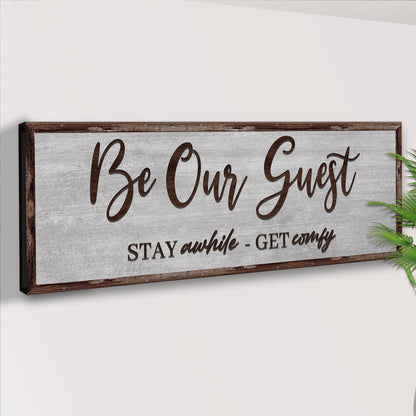 Be Our Guest Stay Awhile canvas on a pale wall beside a palm leaf, angled view
