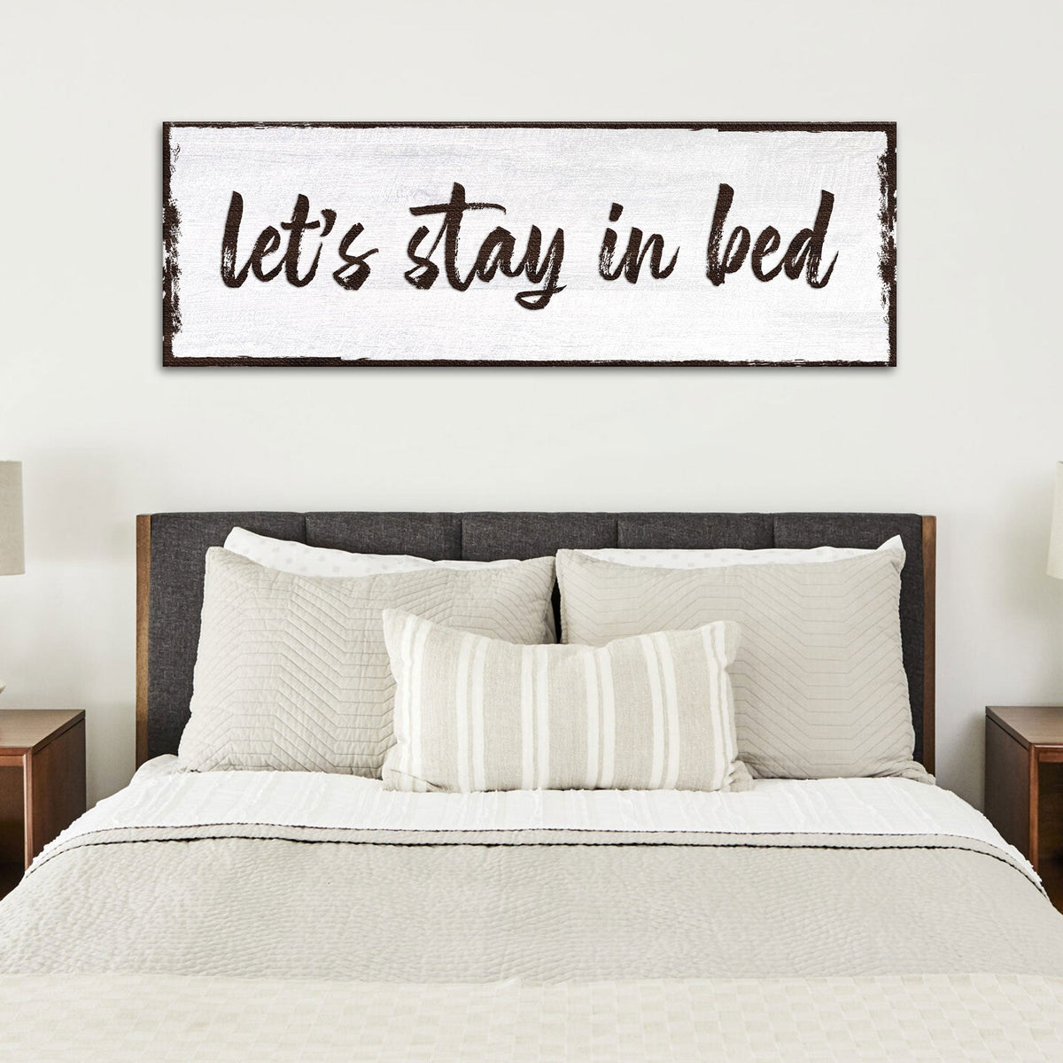 Long white canvas sign with Let's Stay in Bed in dark brush script, above a bed with a dark headboard