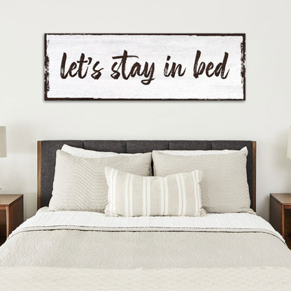 Long white canvas sign with Let's Stay in Bed in dark brush script, above a bed with a dark headboard