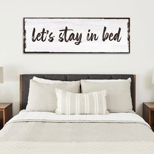 Long white canvas sign with Let's Stay in Bed in dark brush script, above a bed with a dark headboard