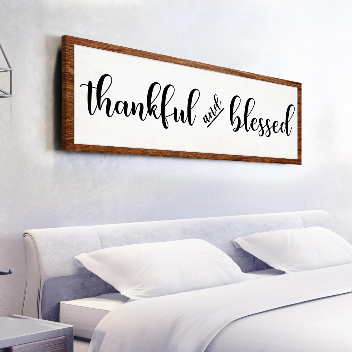 Long white canvas sign reading thankful and blessed in black script with a brown wood-look border, above a bed