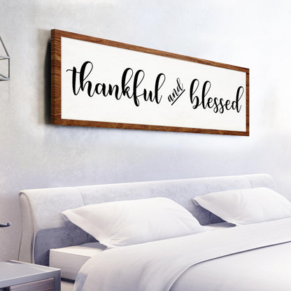 Long white canvas sign reading thankful and blessed in black script with a brown wood-look border, above a bed