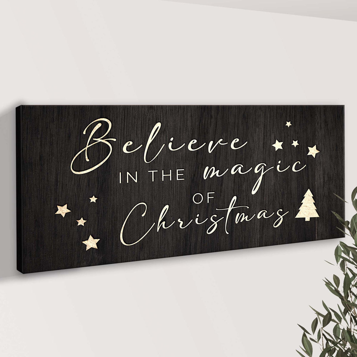 Angled view of the black Believe in the Magic of Christmas canvas sign on a pale wall, showing cream stars and a small tree