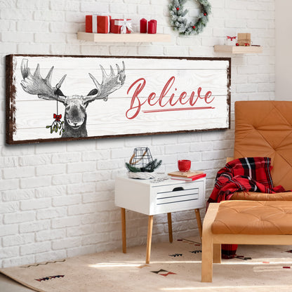Believe Christmas moose canvas sign on a white brick wall beside an orange armchair and a plaid blanket