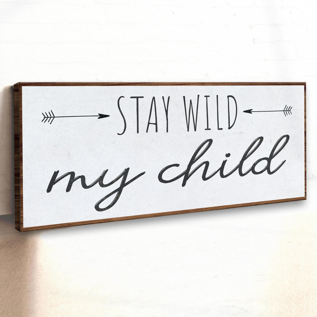 Stay Wild My Child canvas sign seen at an angle on a white wall, showing its brown wood-look border