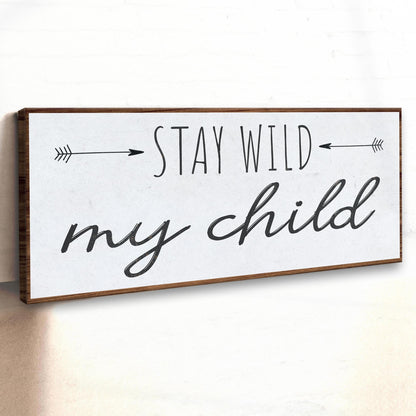 Stay Wild My Child canvas sign seen at an angle on a white wall, showing its brown wood-look border