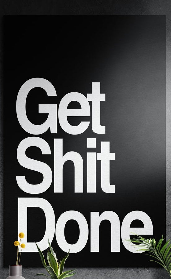 White on black bold Get Sh*t Done brushed aluminum wall art on a dark textured wall above potted plants