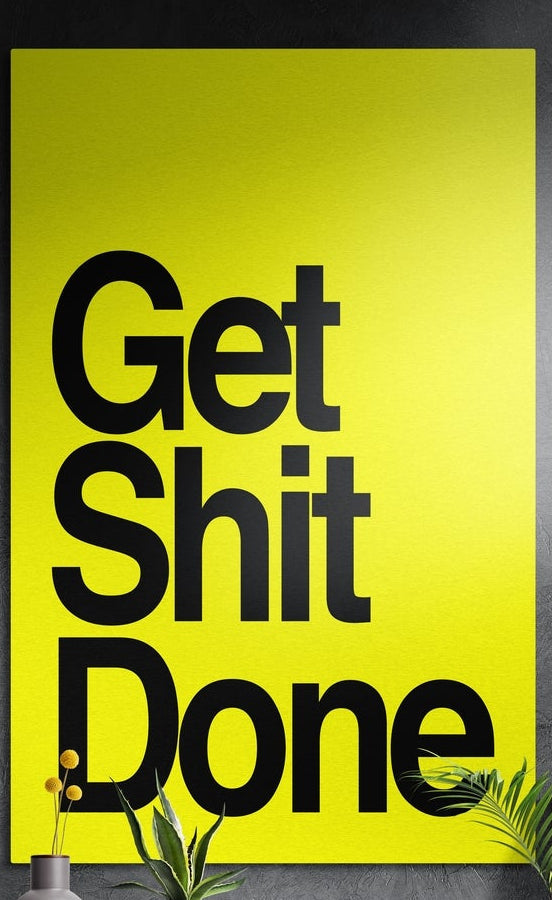 Black on yellow bold Get Sh*t Done brushed aluminum wall art on a dark textured wall above potted plants