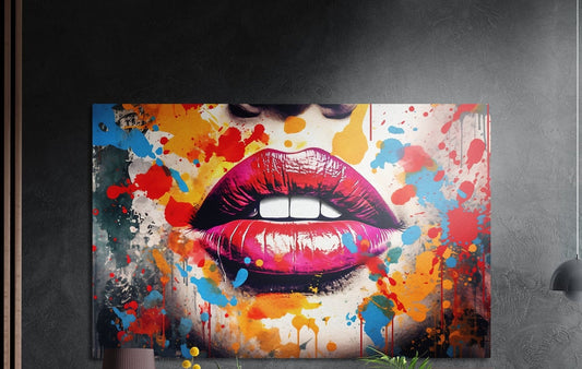 Full pink lips over white teeth amid orange and blue paint splatter, brushed aluminum wall art on a dark wall beside a lamp