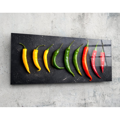 Chili peppers on black slate panoramic glass wall art on a pale concrete wall, angled to show the glossy surface