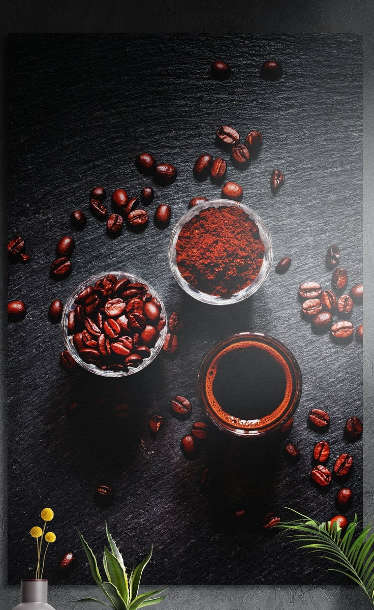 Coffee beans, grounds and a cup on dark slate seen from above, brushed aluminum wall art on a gray wall above yellow flowers