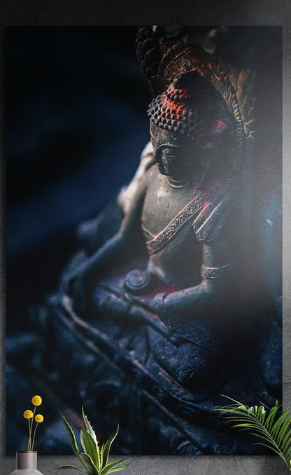 Praying Buddha statue brushed aluminum wall art in dark blue tones on a dark gray wall above potted plants