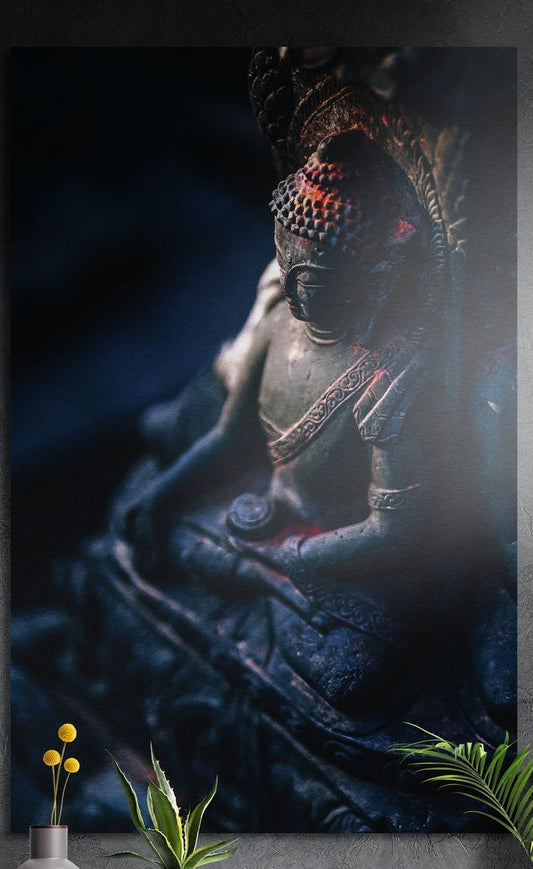 Praying Buddha statue brushed aluminum wall art in dark blue tones on a dark gray wall above potted plants