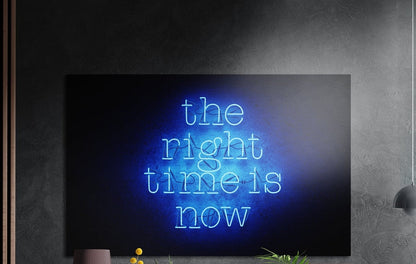 The right time is now blue neon lettering brushed aluminum wall art on a dark textured wall above a lamp and plants