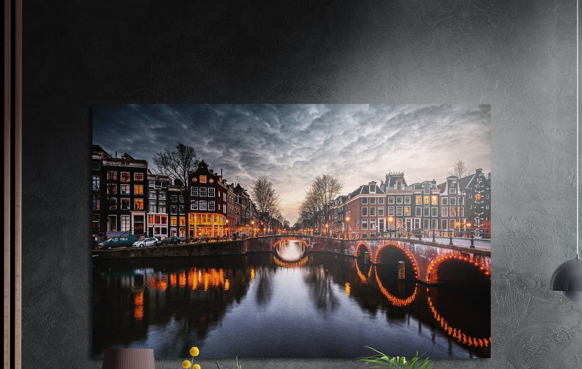 Canal houses and arched bridge at dusk brushed aluminum wall art on a dark textured wall, a lamp and plant tops below