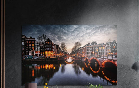 Canal houses and arched bridge at dusk brushed aluminum wall art on a dark textured wall, a lamp and plant tops below