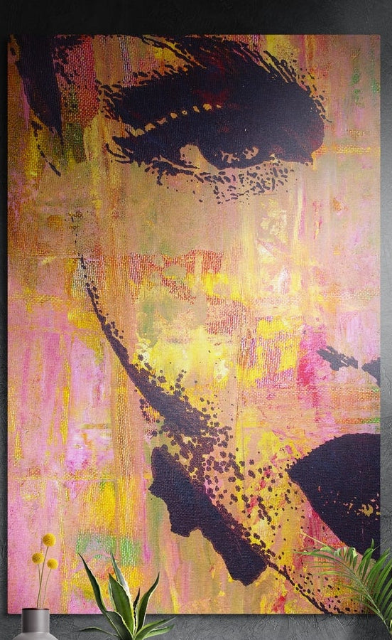 Pink and yellow abstract face brushed aluminum wall art on a dark textured wall above yellow flowers and plants