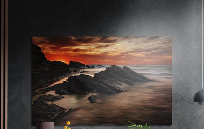 Rocky shore sunset brushed aluminum wall art on a dark gray wall above a sideboard with a vase and plants