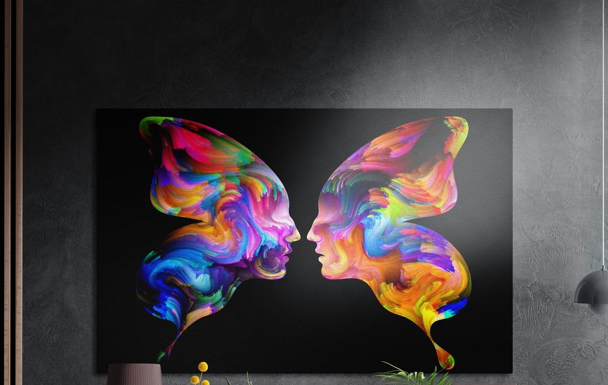 Two rainbow faces in profile forming butterfly wings on black, brushed aluminum wall art on a gray wall above a vase