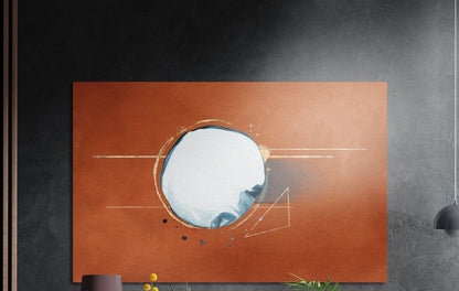 Burnt orange abstract moon circle brushed aluminum wall art on a gray concrete wall with a lamp and plants below