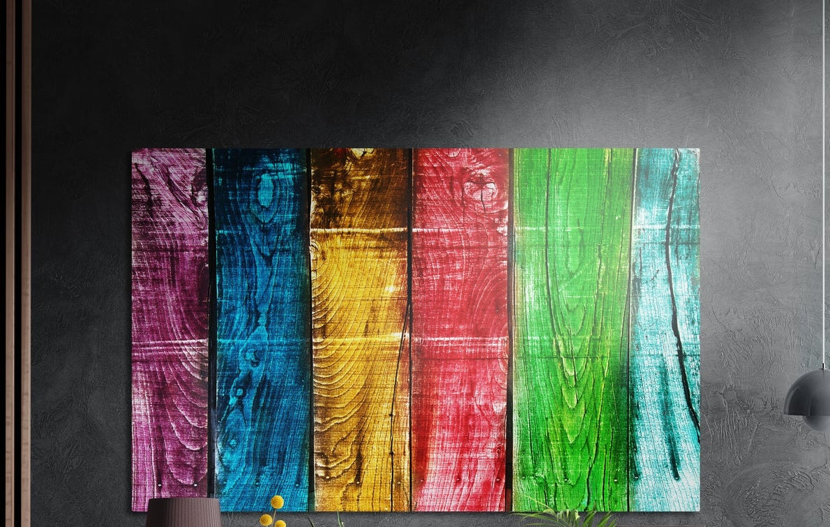 Rainbow wood plank brushed aluminum wall art on a dark textured wall above a lamp and yellow flowers