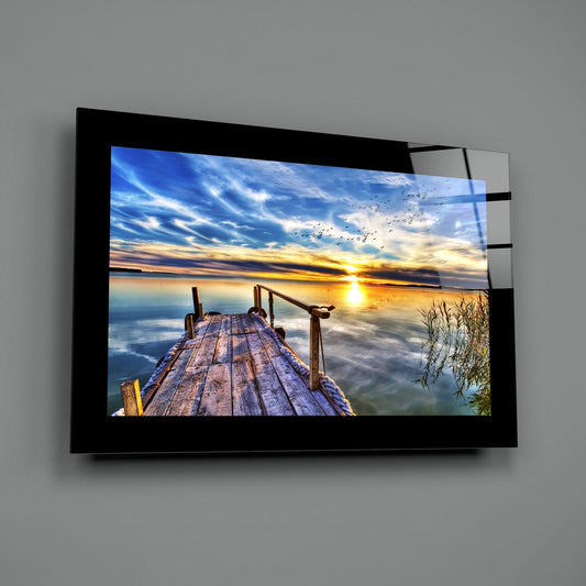Wooden lake pier with reeds at golden sunset glass wall art with a black border hung on a plain gray wall