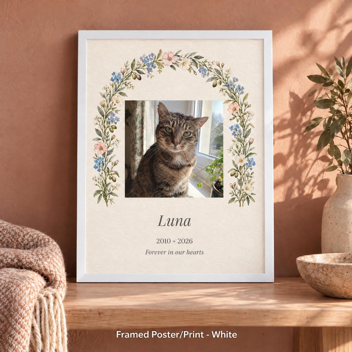 Cat memorial frame in white with a tabby cat photo, name and years, leaning on a bench against a warm wall