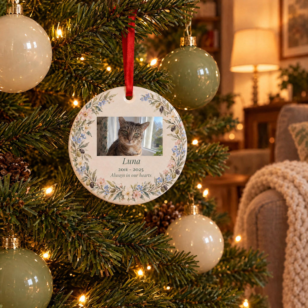 Cat memorial ornament with a tabby cat photo hanging on a lit Christmas tree in a cozy living room