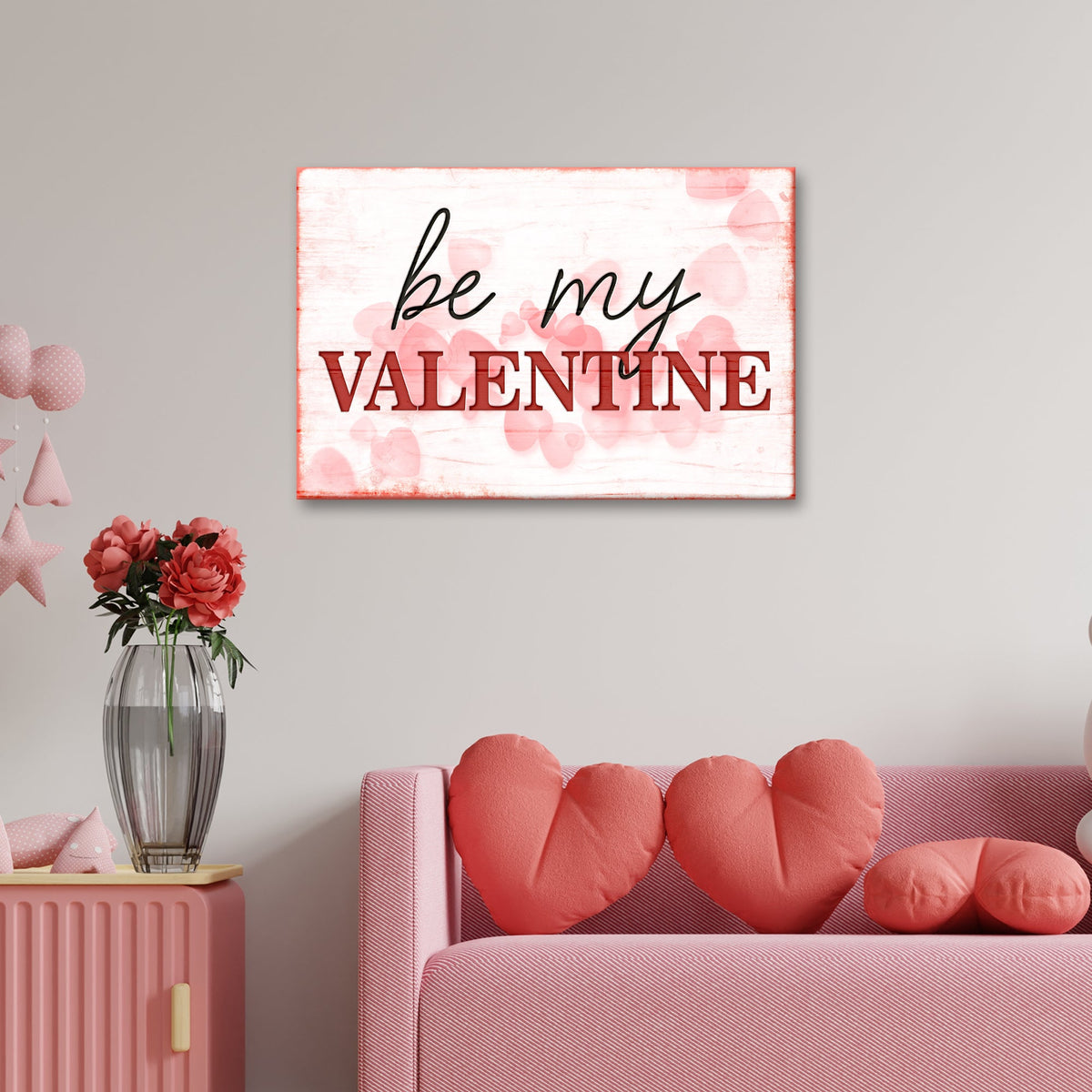 White and pink canvas sign reading Be My Valentine over soft pink hearts, above a pink sofa with heart pillows