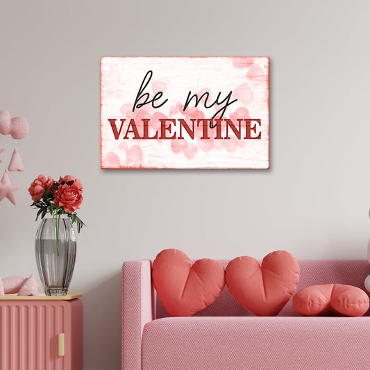 White and pink canvas sign reading Be My Valentine over soft pink hearts, above a pink sofa with heart pillows