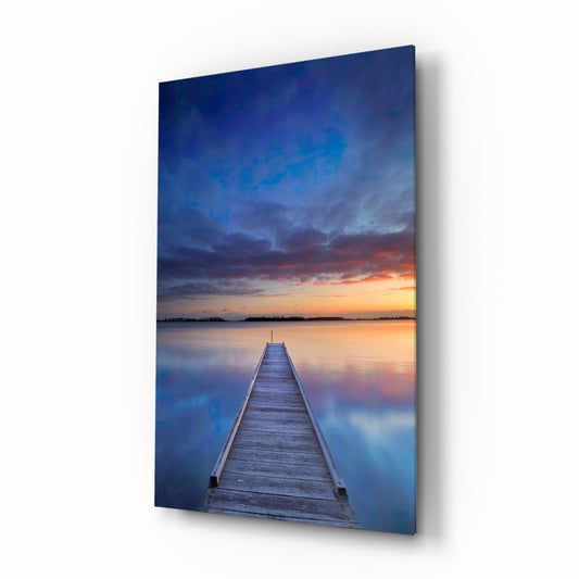 Vertical glass print of a wooden pier stretching into still water under a deep blue sky with an amber sunset
