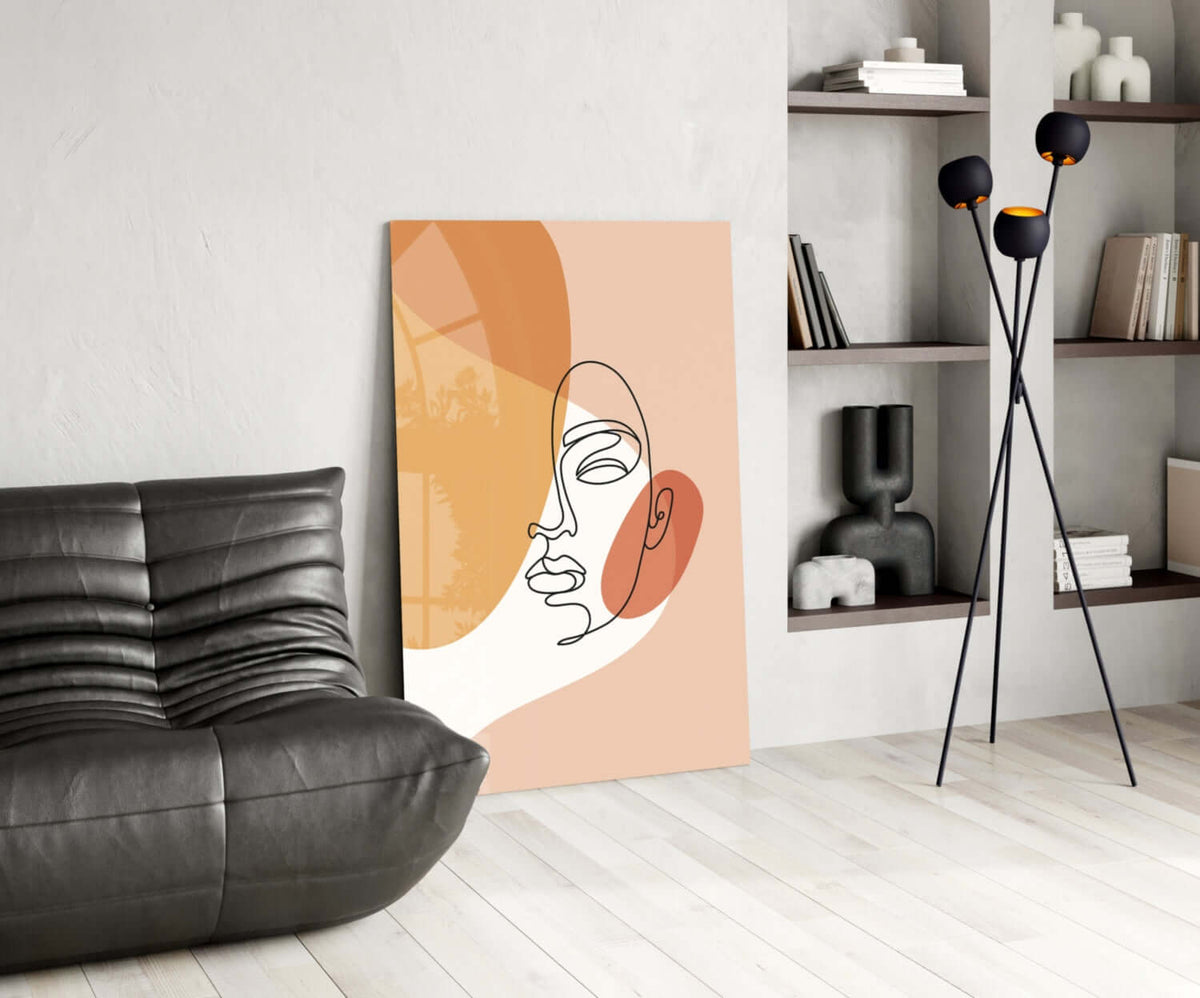 Peach line art face glass wall art leaning on a wall beside a black leather lounge chair and shelves